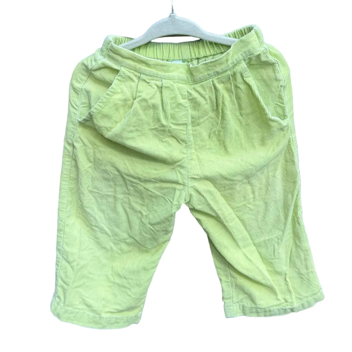 A pair of light yellow-green toddler shorts hanging on a white hanger against a red-brown brick wall with green trees in the background.