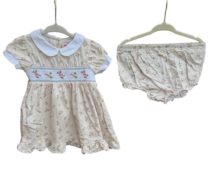 A baby girl's two-piece outfit set consisting of a cream-colored dress with a white Peter Pan collar and a matching floral bloomers, hanging on hangers against a brick wall with trees in the background.