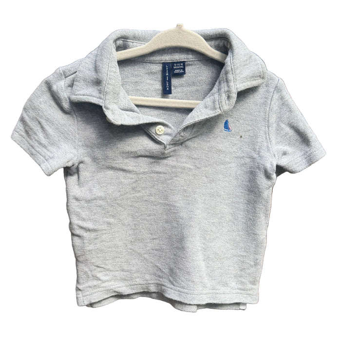 A light gray heather short-sleeve polo shirt for toddlers, featuring a small blue embroidered polo player logo on the left chest, hanging on a white plastic hanger against a red-brown brick wall.