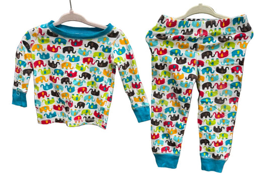 A children's two-piece pajama set hanging on wooden hangers against a brick wall outdoors. The set includes a long-sleeve top and matching pants with a colorful elephant print on a white background and solid blue cuffs and collar.