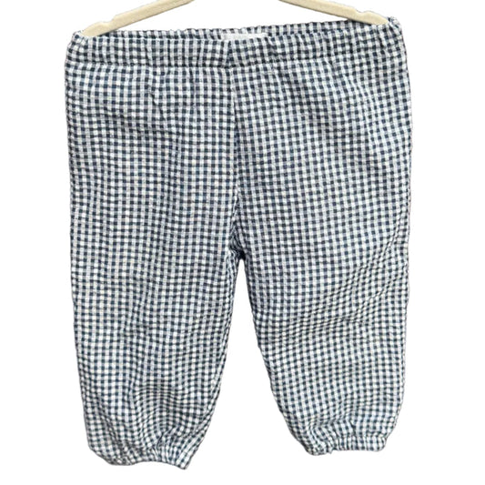 A pair of children's gingham pants in a black and white checkered pattern, hanging on a wooden hanger against a brick wall with green trees in the background.