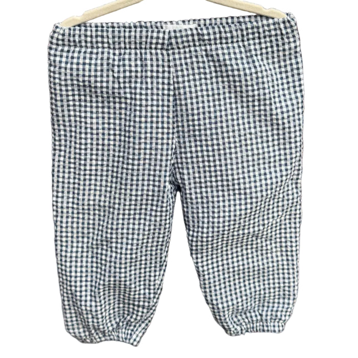 A pair of children's gingham pants in a black and white checkered pattern, hanging on a wooden hanger against a brick wall with green trees in the background.