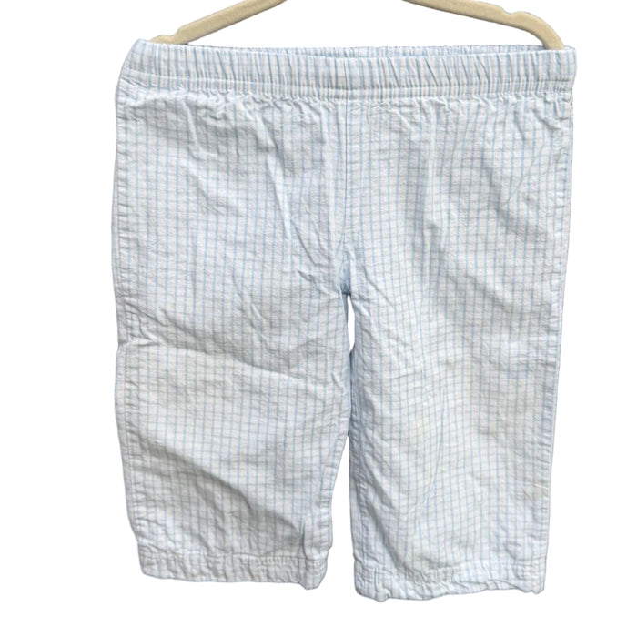 Light blue and white checkered shorts hanging on a beige hanger against a red-brown brick wall with green foliage in the background.