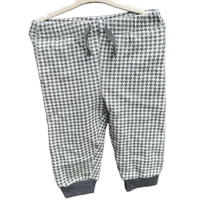 A pair of children's jogger pants with a black and white houndstooth pattern, hanging on a white plastic hanger against a red-brown brick wall.