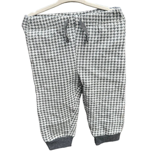A pair of children's jogger pants with a black and white houndstooth pattern, hanging on a white plastic hanger against a red-brown brick wall.