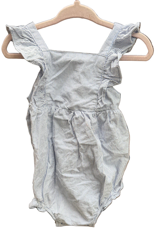 A baby romper with light blue and white vertical pinstripes, featuring ruffled shoulder straps and a smocked waist, hanging on a wooden hanger against a brick wall.