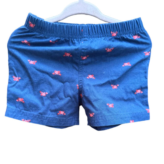 Blue children's shorts with a pink lobster print, hanging on a white plastic hanger against a brick wall background.