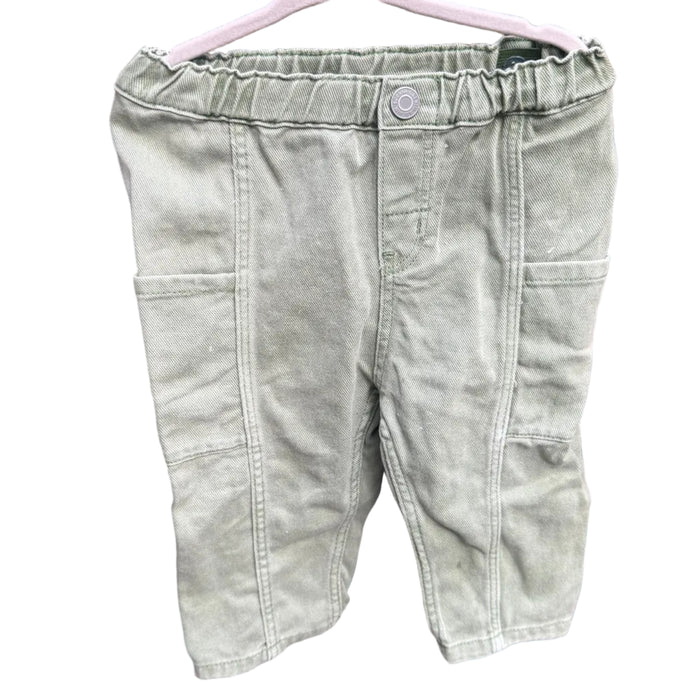 A pair of light sage green denim shorts hanging on a white plastic hanger against a red-brown brick wall.