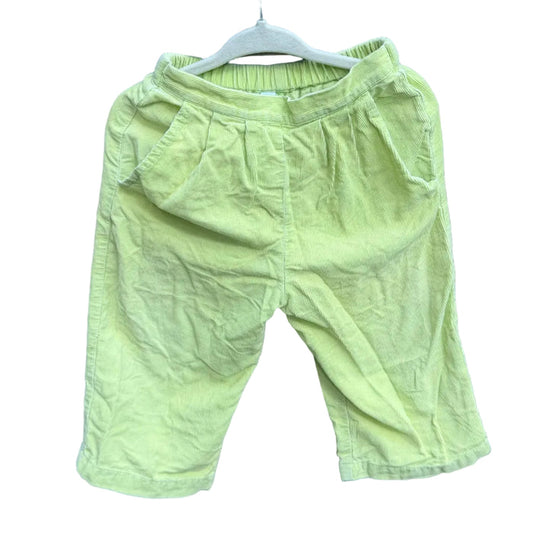 A pair of light yellow-green toddler shorts hanging on a white hanger against a red-brown brick wall with green trees in the background.