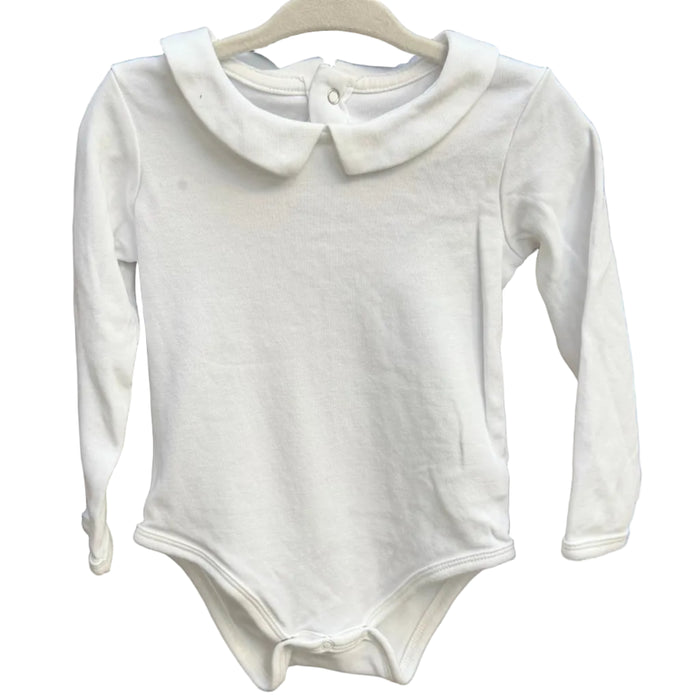 A white long-sleeve baby bodysuit with a Peter Pan collar hangs on a wooden hanger against a rustic brick wall.