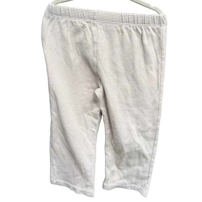 A pair of light beige or off-white cropped pants hanging on a white plastic hanger against a red-brown brick wall with green foliage visible above.