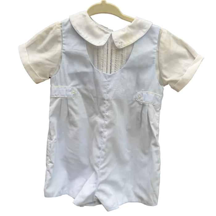 A baby boy's short-sleeve white romper with a Peter Pan collar and buttoned chest pockets, hanging on a wooden hanger against a red-brown brick wall.
