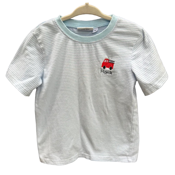 A light blue and white striped short-sleeve t-shirt with a light blue collar, featuring a small red fire truck graphic and the name 'Frank' printed on the chest, hanging on a wooden hanger against a brick wall.
