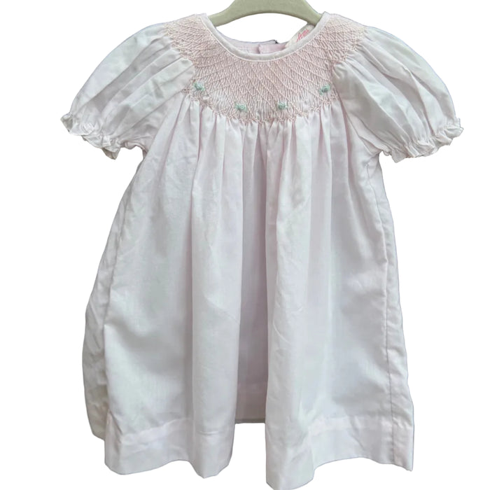 A light pink baby smocked dress with short puff sleeves hanging on a white plastic hanger against a brown brick wall with green foliage in the background.