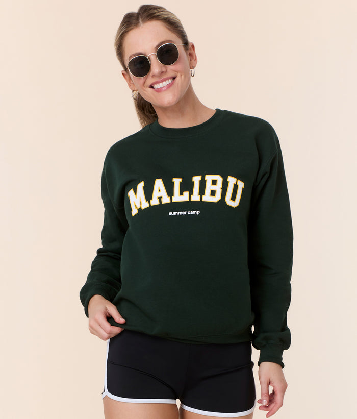 Model wearing the Mindy Sweatshirt in forest green cotton by Andie Swim with 'Malibu summer camp' varsity lettering, perfect for reliving summer camp days.