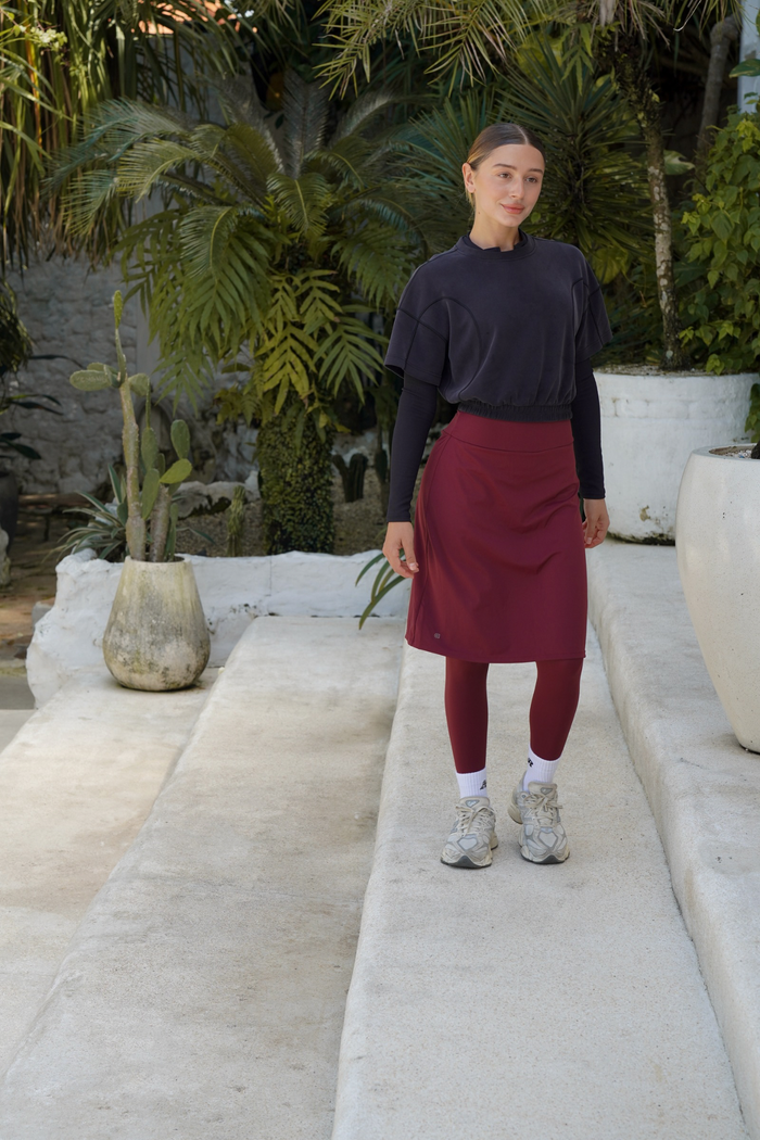 Person wearing a black top and red skirt standing on a pathway with plants in the background. Modest sport skirt with integrated legging in muted wine, designed for modest activewear and comfortable movement for women.