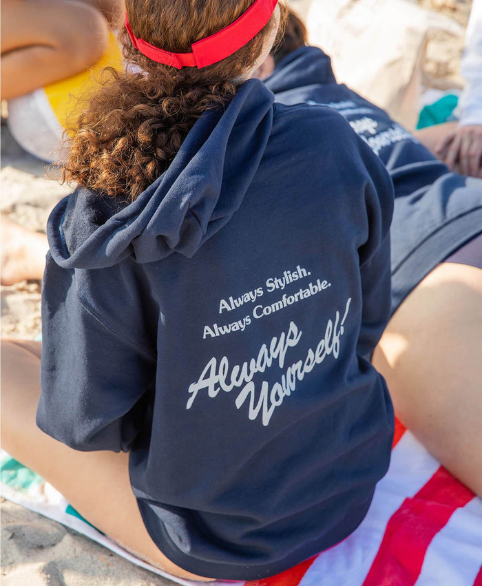 Model wearing the Always Full Zip Hoodie Sweatshirt in Navy, sitting on a beach with back facing the camera. The hoodie features the Andie motto: 'Always Stylish. Always Comfortable. Always Yourself!' in bold white print