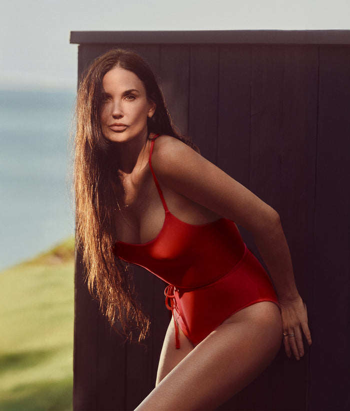 A woman posing confidently outdoors in The Fiji One Piece swimsuit in Cherry Red. The eco-nylon suit features a scoop neckline, adjustable straps, and a cinched waist detail for a flattering classic fit, perfect for a sophisticated beach or poolside look.